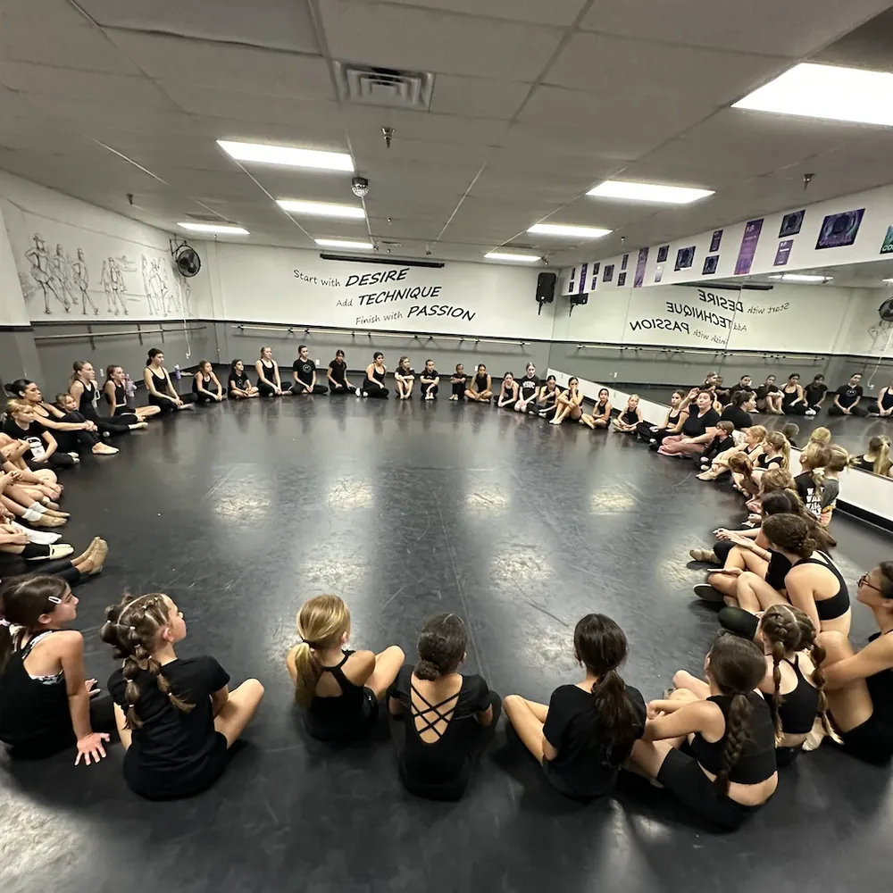 MSDC dancers sitting in a circle across the studio floor, beneath the studio's motto on the wall